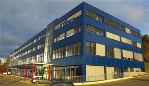 An aerial image of a one story building, lined with windows and with a sign that reads Cohu. There is a defined, paved path to enter lined with shrubs and cars in the foreground of the parking lot.