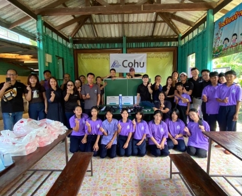 A group of 32 children and adults smiling at the camera making hearts with their fingers. The children are wearing purple shorts and a sign reading 'Cohu' is behind them in an open air building