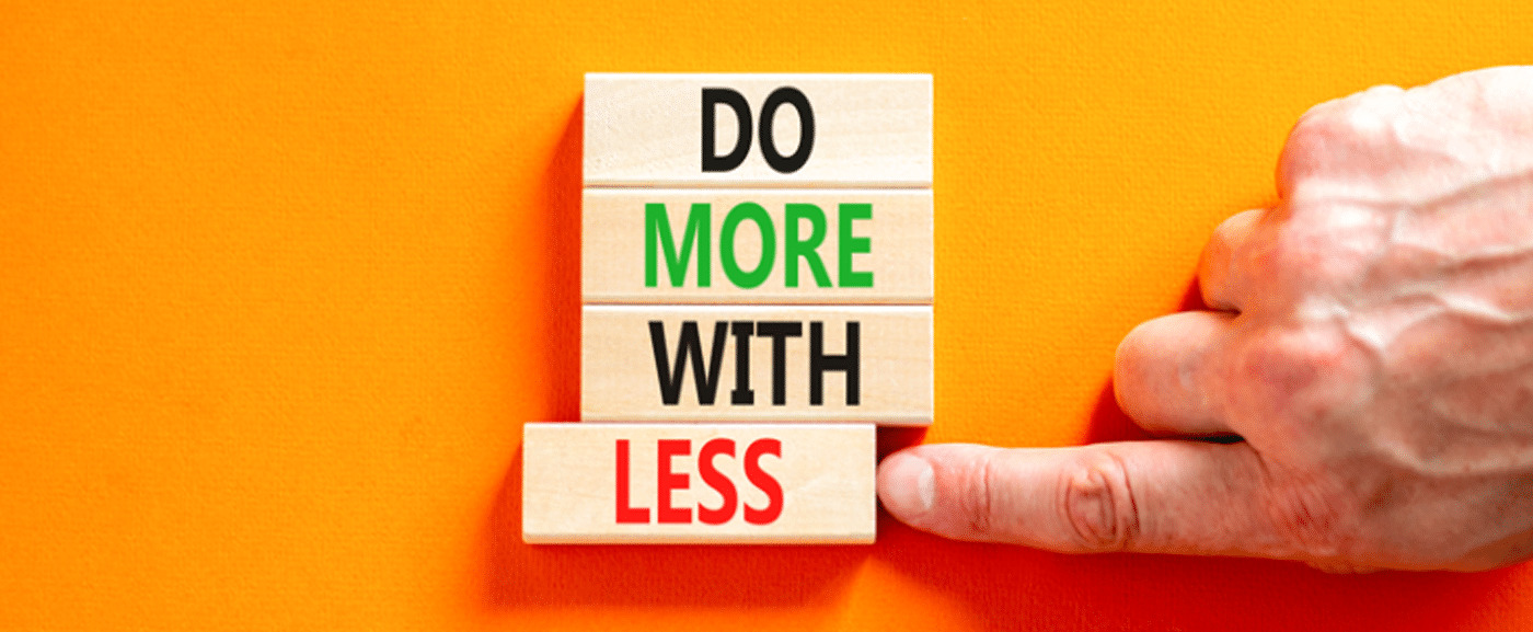 An orange background with four wooden blocks arranged vertically. Each one reads one word 'Do' 'More' 'With' 'Less' and there is a hand pushing the last block 'Less' out of alignment with the others