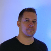 Headshot of a man looking at the camera wearing a black sweater and a blue light projected onto a white wall for the background